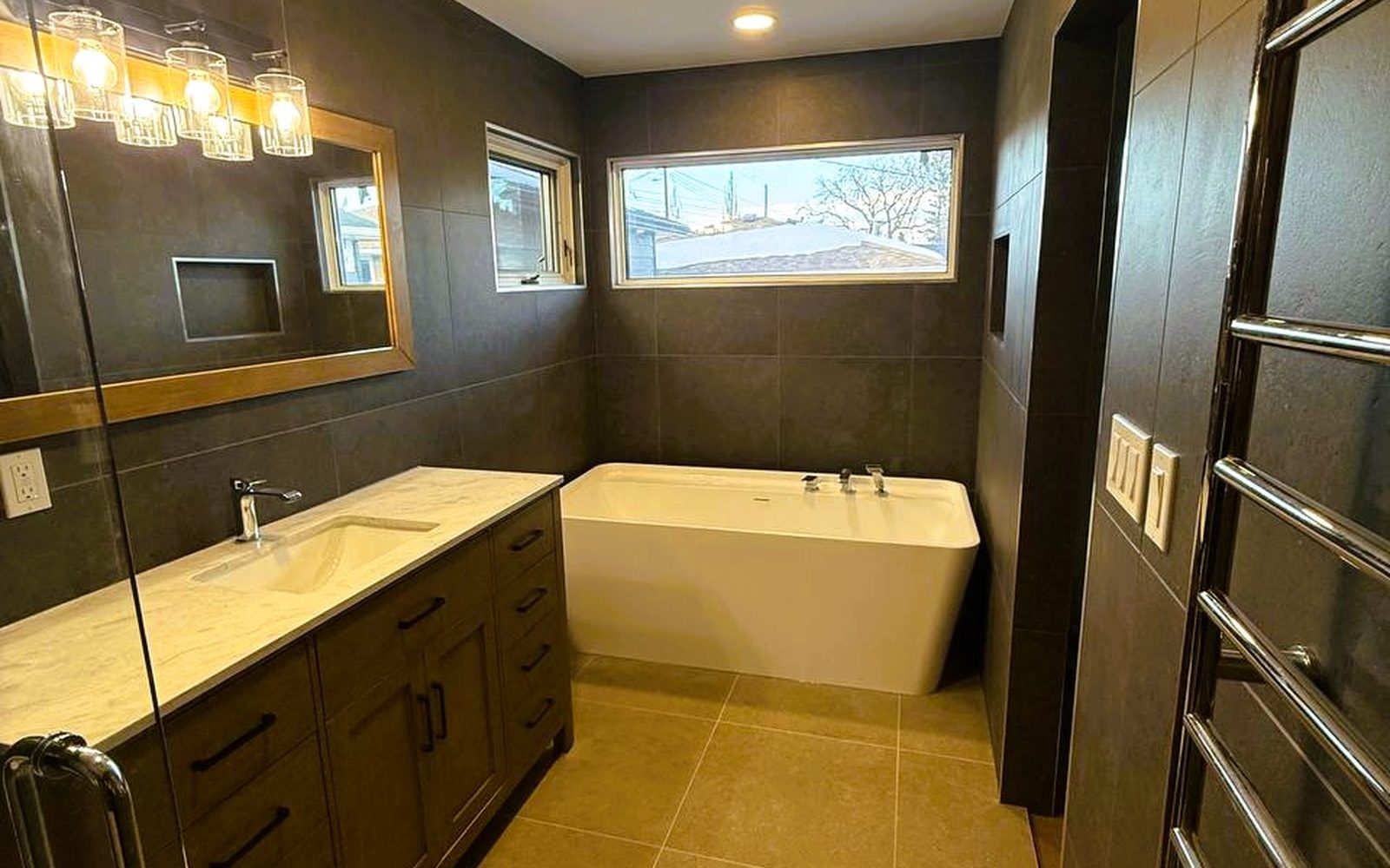 Finished bathroom built by Lions Group: long wood vanity and a freestanding tub under a wide window, in large-format brown stone tile
